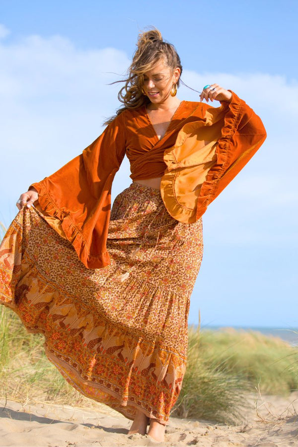 model styling one of our Velvet Venus Wrap Top in marmalade yellow. The iconic mustard gold warm tone top in a soft shimmering velvet. Featuring huge bell sleeves with a frill edge. Shown tied at the front inspired by 70's bohemia styles. By All About Audrey